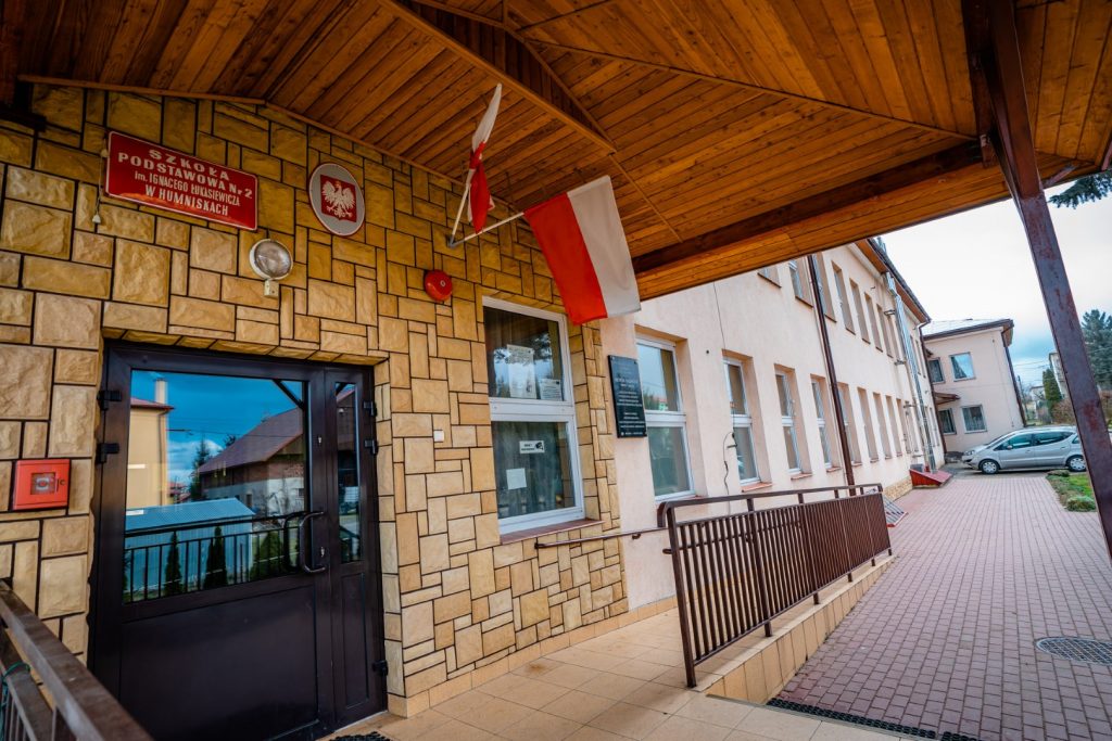 Oszklone drzwi prowadzące do szkoły. Nad wejściem czerwona tablica z białymi literami oraz godło Polski