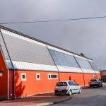 Bydynek hali sportowej z czerwoną elewacją. Przed halą trzy zaparkowane samochody