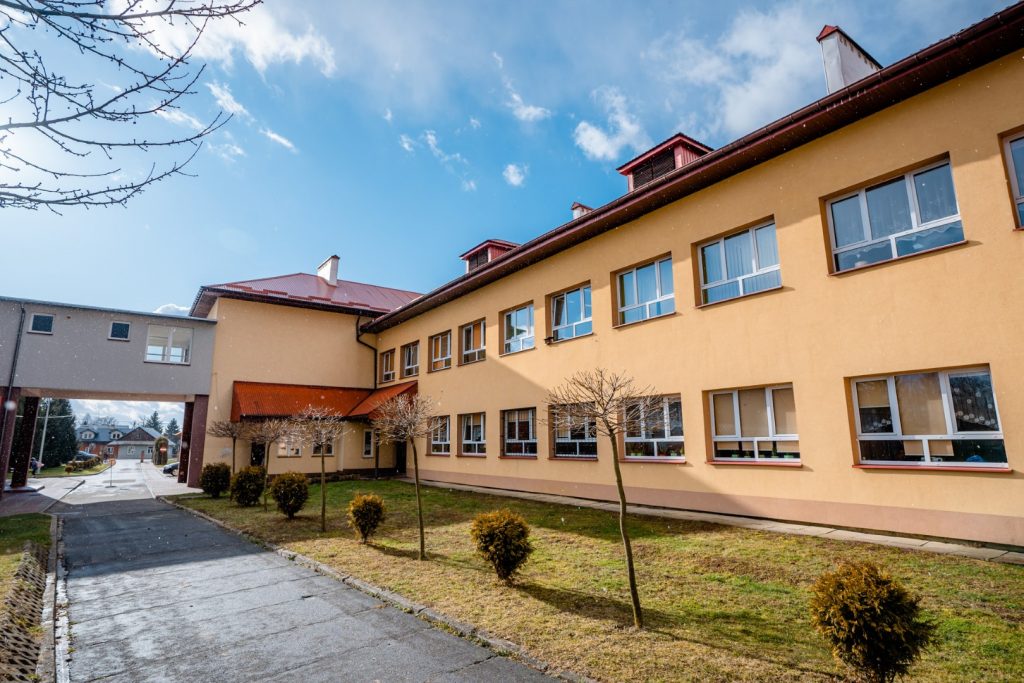 Dwukondygnacyjny budynek szkolny z elweacją w kolorze żółtym. Przed szkołą trawnik, krzewy i drzewka