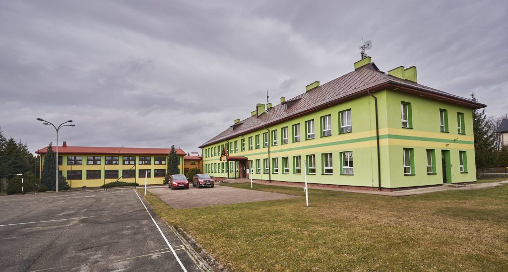 Budynek szkoły z zieloną elewacją i brązowym dachem. Przed szkołą stoją dwa samochody