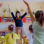 Kobieta w niebieskiej bluzce podnosi ręce . Przed nia grupa dzieci, która naśladuje jej ruchy