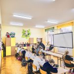 Dzieci siedzą w ławkach i piszą w zeszytach. N aśrodku tablica interaktywna