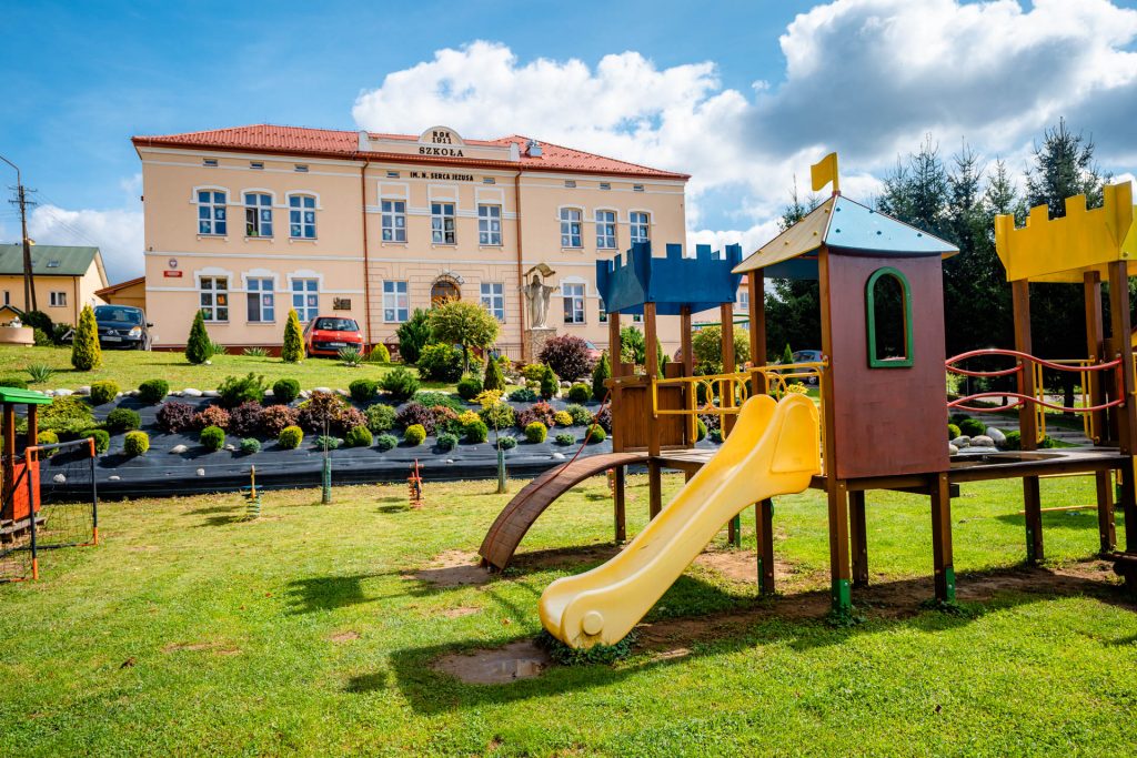 Plac zabaw wśród zieleni. W głębi różowy budynek ze spadzistym dachem