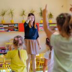 Kobieta w niebieskiej bluzce wskazuje palcem na dziecko. Przed nia grupa dzieci, która naśladuje jej ruchy