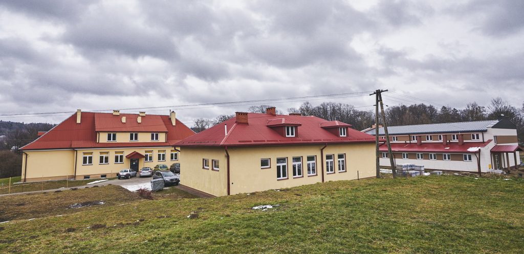 Budynki szkolne z czerwonymi dachami. Ściany elewacji pomalowane na żółto. Z boku biało-brązowy budynek z blaszanym dachem