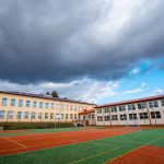 Dwukondygnacyjne budynki szkolne ze spadzistymi dachami. Przed budynkami boisko sportowe ze sztuczną nawierzchnią