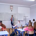 Klasa szkolna, uczniowie siedzą w ławkach. Nauczycielka stoi przy tablicy. Na ścianie godło Polski i krzyż