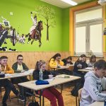 Dzieci siedzą w ławkach w klasie. Na ścianie koloru zielonego namalowana scena bitewna