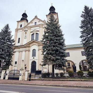 Zabytkowy kościół z dwoma wieżami. Elewacja koloru beżowego. Przed kościołem rosną drzewa