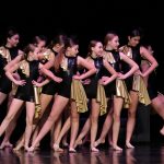 Dziewczyny w czarnych, błyszczących kombinezonach ze złotymi falbankami tańczą na scenie