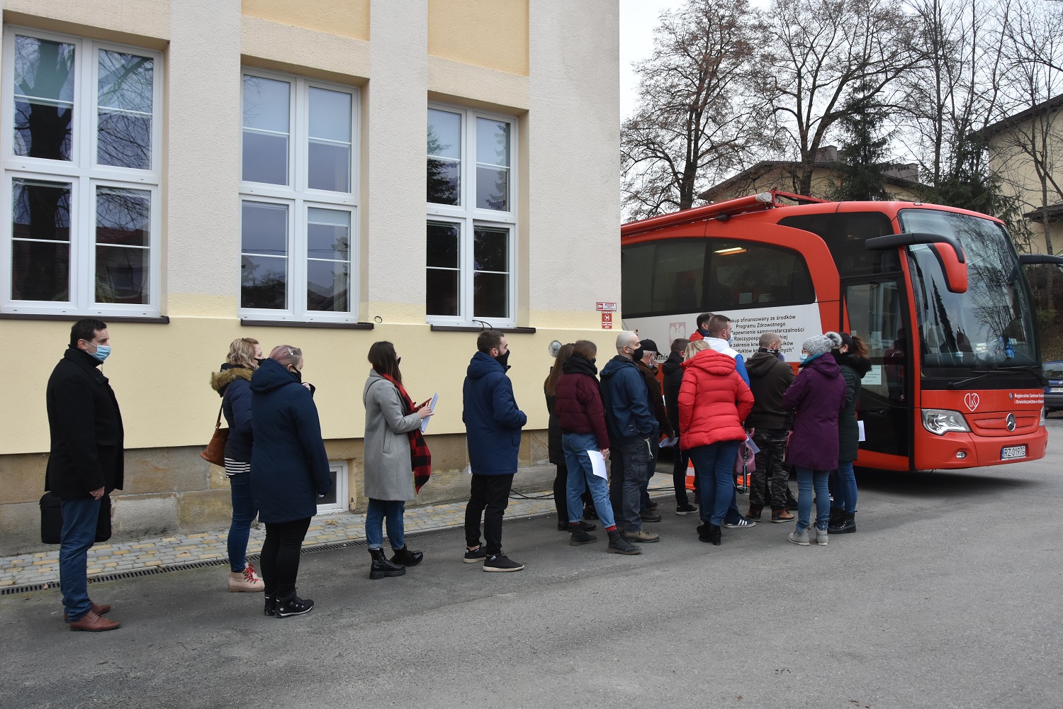 Kolejka kilkudziesięciu ludzi (mężczyzn i kobiet) oczekujących na pobranie krwi w specjalnym do tego przygotowanym autobusie koloru czerwonego.