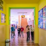 Grupa dzieci w maseczkach na korytarzu w szkole. Ściany na korytarzu są koloru zielonego, korytarz wyłożony płytkami brązowymi.