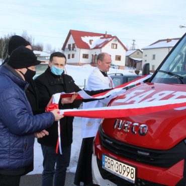 Trzech mężczyzn, w tym ksiądz przecina czerwono - białą wstęgę na samochodzie pożarniczym.