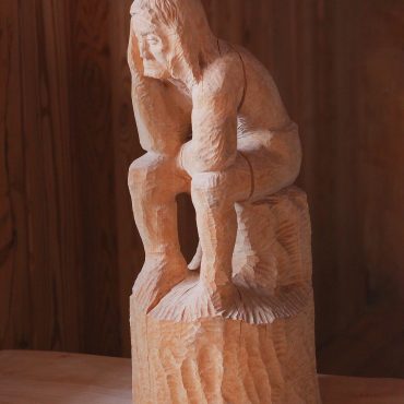 Figurka z drewna przedstawiająca mężczyznę siedzącego na pniu z trzymającymi ręce na głowie.