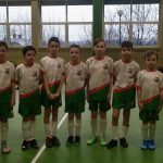 Grupa kilkudziesięciu chłopców w różnokolorowych strojach piłkarskich, stoją na sali gimnastycznej. Obok nich stoi dwóch trenerów.