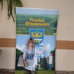 Dziewczynka w krótkich jasnych włosach stoi przed banerem powiatu brzozowskiego i trzyma w rękach dyplom i gadżety promocyjne, tj. jasny notatnik.