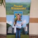 Dziewczynka w jasnych włosach z opaską na głowie stoi przed banerem powiatu brzozowskiego i trzyma w rękach dyplom i puchar.