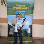 Chłopczyk w okularach stoi przed banerem powiatu brzozowskiego i trzyma w rękach dyplom i gadżety promocyjne, tj. jasny zeszyt.