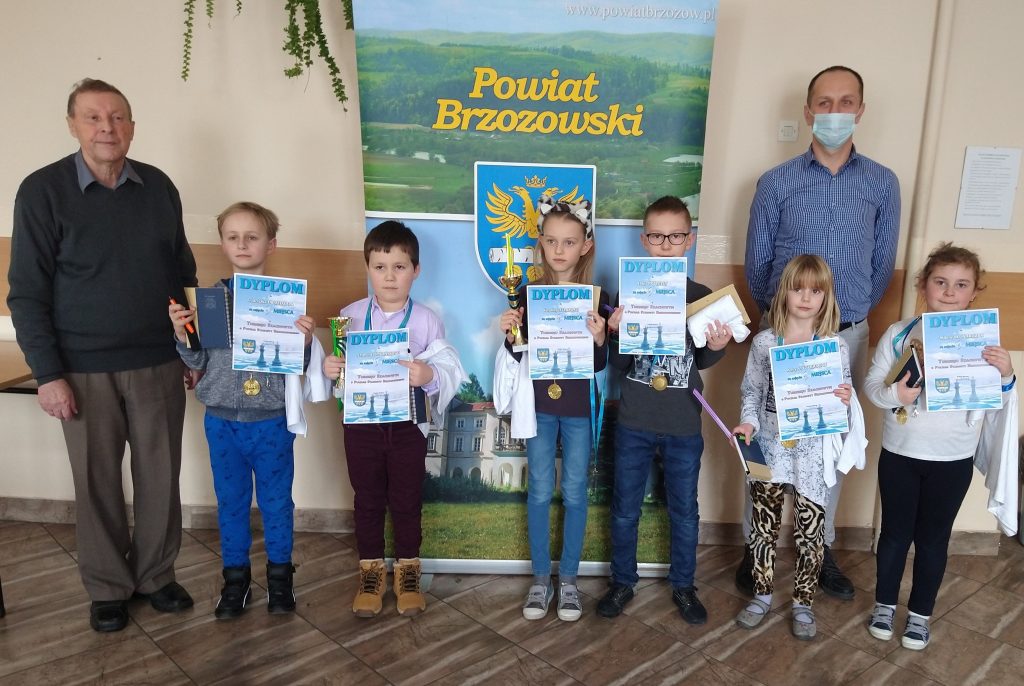 Grupa dzieci oraz dwóch opiekunów stoją przed banerem Powiatu Brzozowskiego. Dzieci trzymają puchary, dyplomy i upominki. 