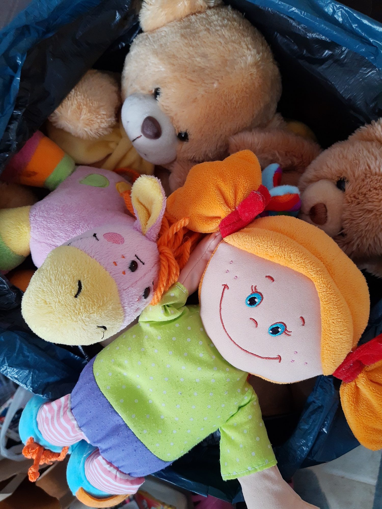 Pluszaki dla dzieci. Wśród nich: szmaciana lalka z pomarańczowymi włosami upiętymi w dwa kucyki. Obok pluszowy osiołek koloru różowego oraz dwa brązowe pluszowe misie.