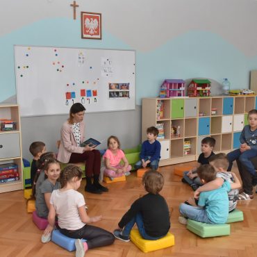 Dzieci w sali zajęć siedzą na podłodze i słuchają nauczycielki która czyta książkę.