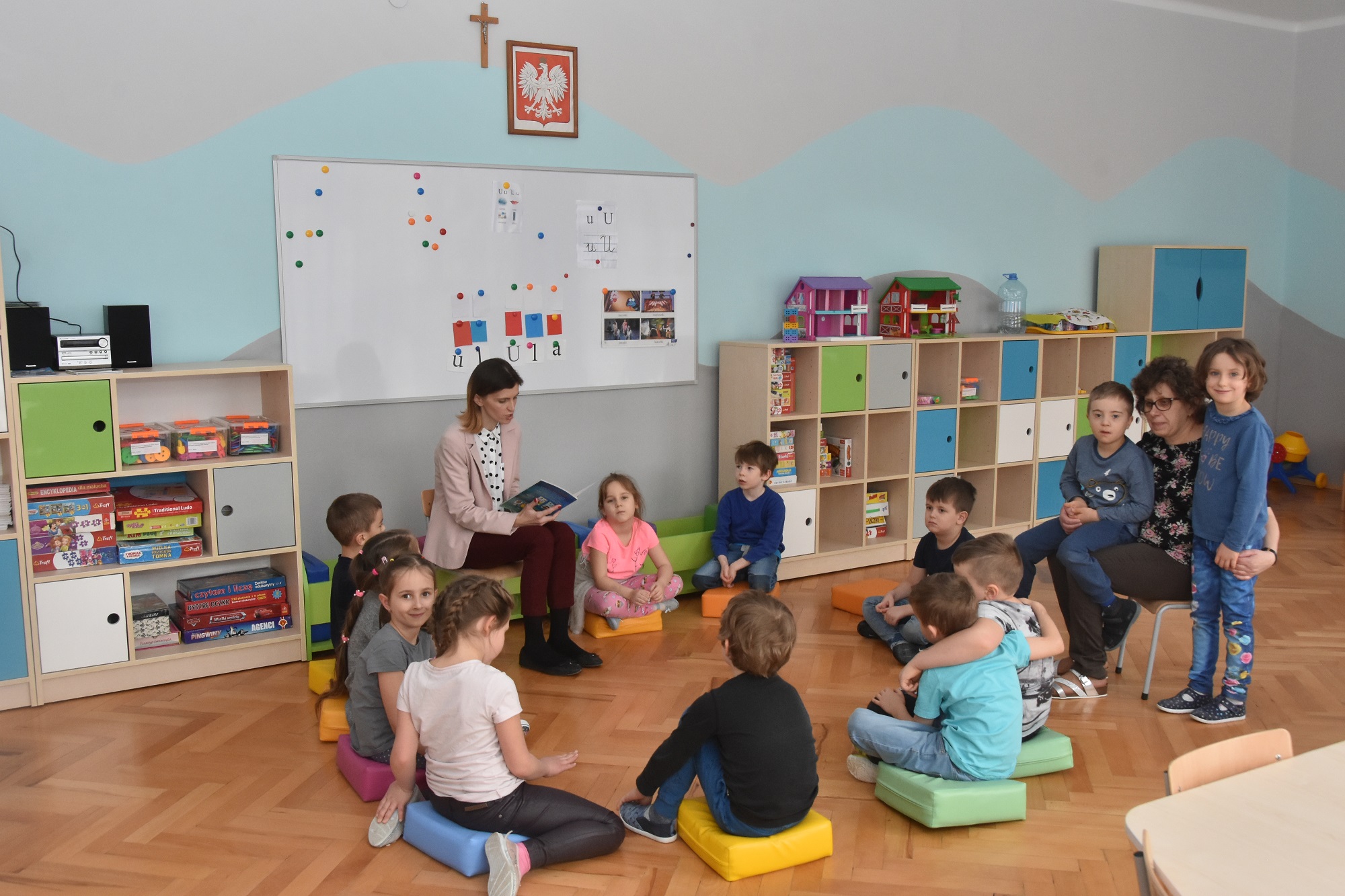 Dzieci w sali zajęć siedzą na podłodze i słuchają nauczycielki która czyta książkę.