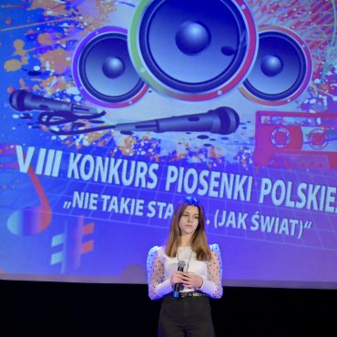 Dziewczyna z ciemnymi włosami, w białej bluzce i ciemnych spodniach śpiewa na scenie. W rękach trzyma mikrofon. Za nią na ekranie wyświetlone logo konkursu.