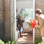 Starsza kobieta stoi w drzwiach, młoda kobieta stoi przed drzwami i trzyma w rękach bukiet kwiatów. Obok rosną zielone krzaki.