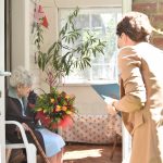 Starsza kobieta siedzi na krześle na ganku w domu, przed drzwiami stoi młoda kobieta i przekazuje bukiet kwiatów.
