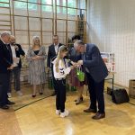 Mała dziewczynka odbiera nagrodę od mężczyzny w garniturze na sali gimnastycznej.