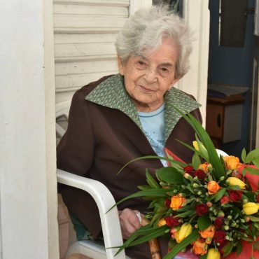 Starsza kobieta siedzi na białym krześle, w rękach trzyma bukiet kolorowych kwiatów.