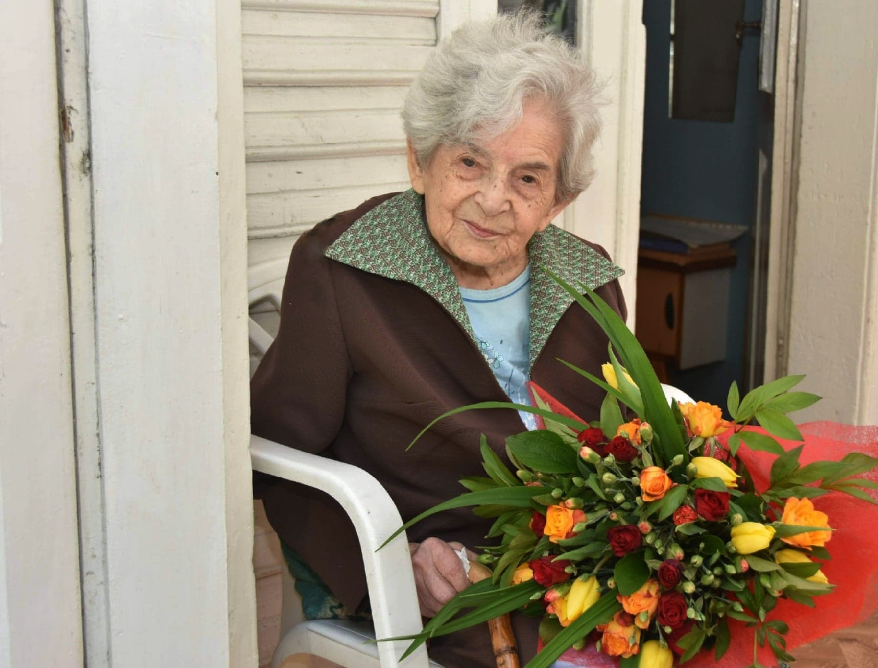 Starsza kobieta siedzi na białym krześle, w rękach trzyma bukiet kolorowych kwiatów.