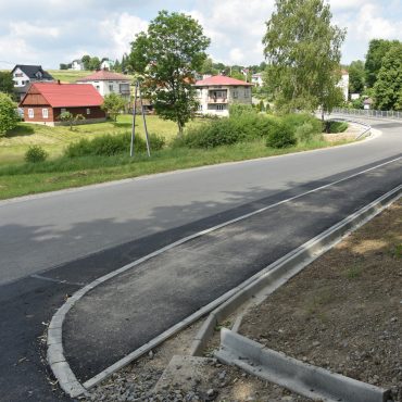Nowy chodnik wzdłuż drogi asfaltowej. Po lewej stronie tereny zabudowane. .