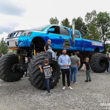 Ogromny samochód monster truck koloru niebieskiego stoi na placu kamienistym. Z boku ogląda go grupa osób.
