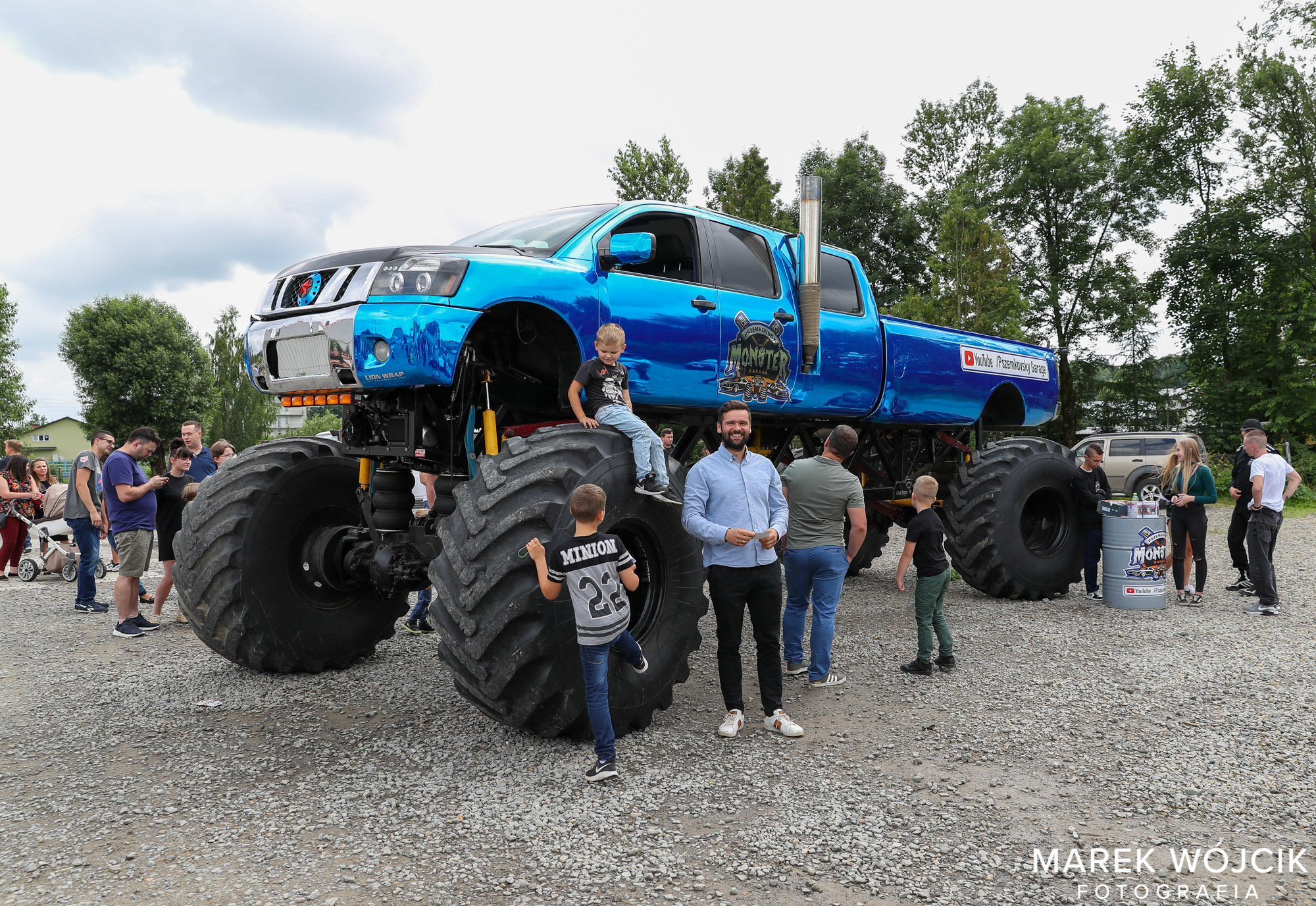 Ogromny samochód monster truck koloru niebieskiego stoi na placu kamienistym. Z boku ogląda go grupa osób.