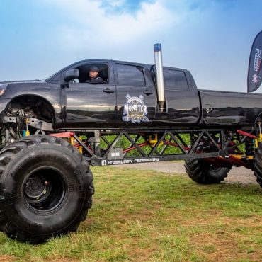 Ogromny samochód monster truck koloru czarnego stoi na trawniku. Za nim wiszą flagi.
