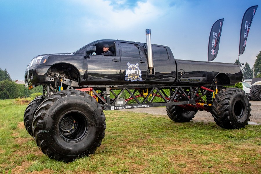 Ogromny samochód monster truck koloru czarnego stoi na trawniku. Za nim wiszą flagi.