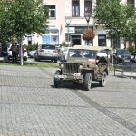 Stary wojskowy samochód jadący po rynku miejskim. W tle zaparkowane samochody.