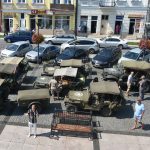 Stare wojskowe smaochody zaparkowane na rynku. Obok nich przechadzają się osoby w różnym wieku.