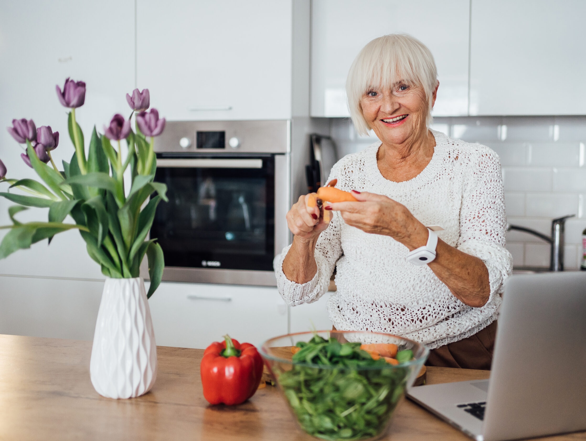Starsza kobieta siedząca przy stole w kuchni. Na stole laptop, kwiaty i warzywa.