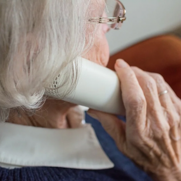 Starsza kobieta z siwymi włosami trzyma słuchawkę przy uchu . Siedzi w domu na krześle.
