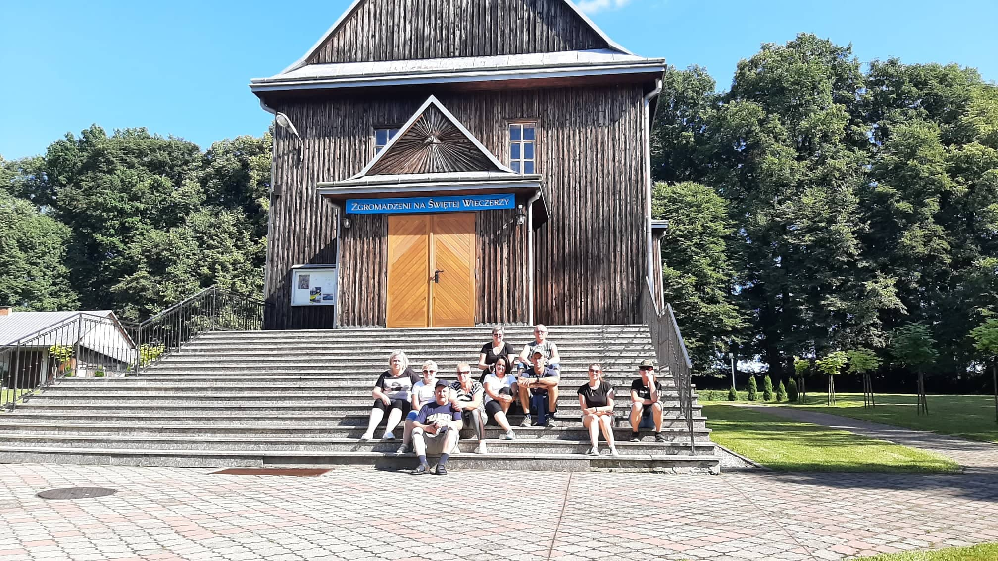 Grupa osób siedzi na schodach do drewnianego kościoła. Obok niego rosną drzewa liściaste.