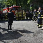 Grupa osób ubrana w stroje strażackie stoi na naradzie w lesie, za nimi stoją samochody pożarnicze.