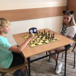 Dziewczynka i chłopiec w jasnych włosach siedzą przy stoliku w pomieszczeniu szkolnym i grają w szachy.