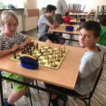 Grupa dzieci siedzą przy stolikach w pomieszczeniu szkolnym i grają w szachy.