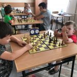 Grupa dzieci siedzi przy stolikach w pomieszczeniu szkolnym i grają w szachy.