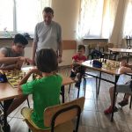 Grupa dzieci siedzą przy stolikach w pomieszczeniu szkolnym grają w szachy. Obserwują ich straszy mężczyzna.