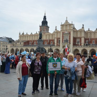 Grupa osób starszych z transparentem Gmina Brzozów, pozuje do zdjęcia na tle kamieniczek i parady w Krakowie. Za nimi panie trzymają balony, inni ubrani są w jednakowe stroje. .