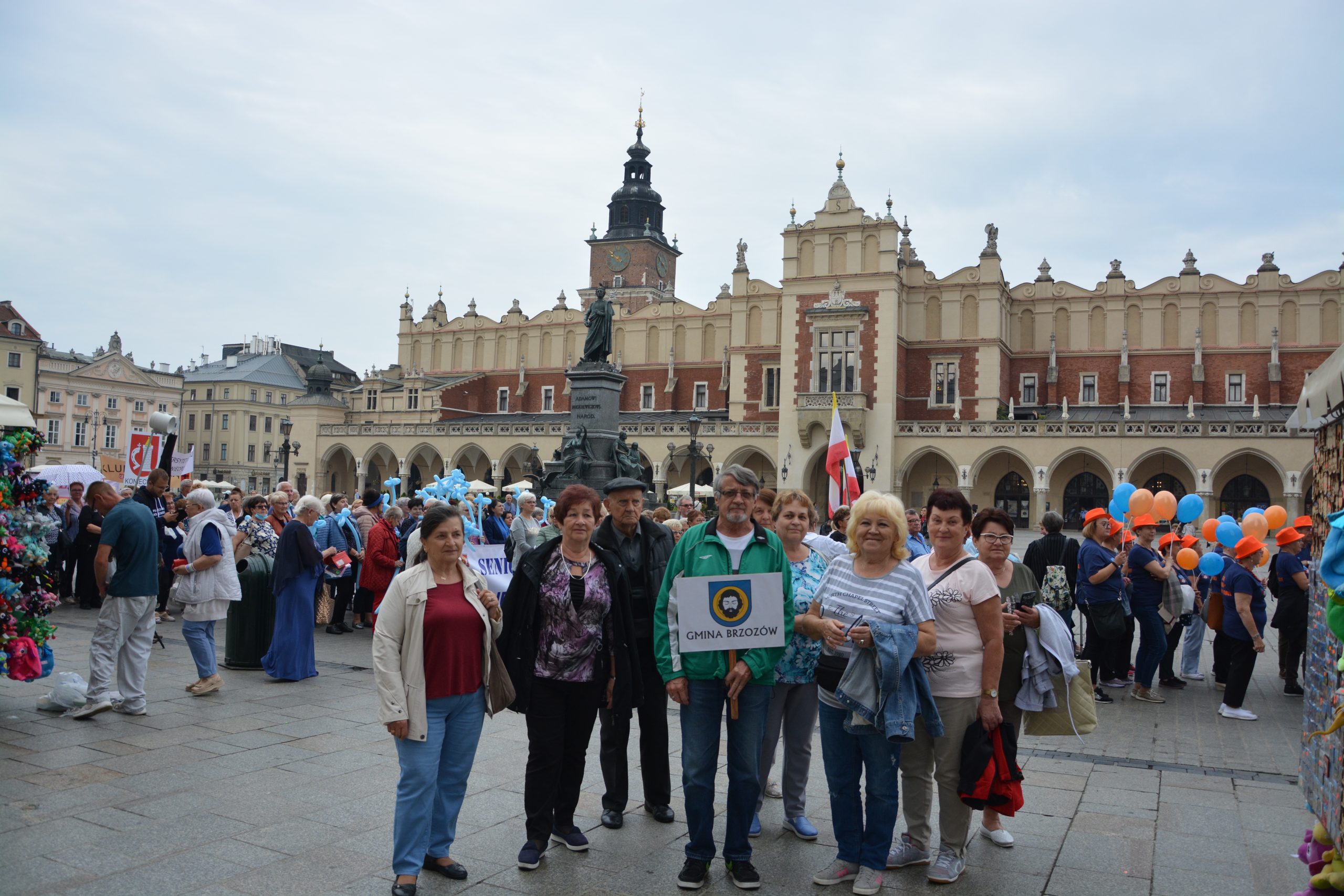 Grupa osób starszych z transparentem Gmina Brzozów, pozuje do zdjęcia na tle kamieniczek i parady w Krakowie. Za nimi panie trzymają balony, inni ubrani są w jednakowe stroje. .