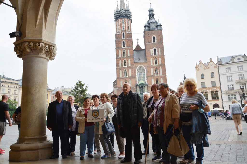 Grupa osób pozuje do zdjęcia na tle ogromengo kościoła, wyłożonego cegłami, Kobieta w centrum zdjęcia trzyma w ręce tabliczkę z napisem Gmina Brzozów. 
